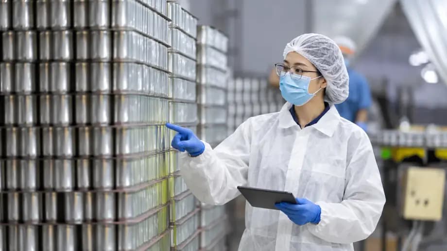 A woman in a white coverall and a face mask pointing at stacked metal cans, a tablet in her other hand